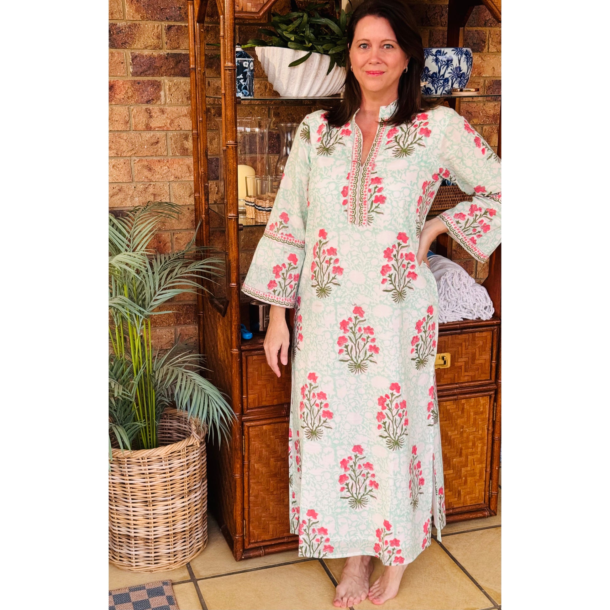 Woman wearing a floral dress standing in a room with wooden furniture and plants.