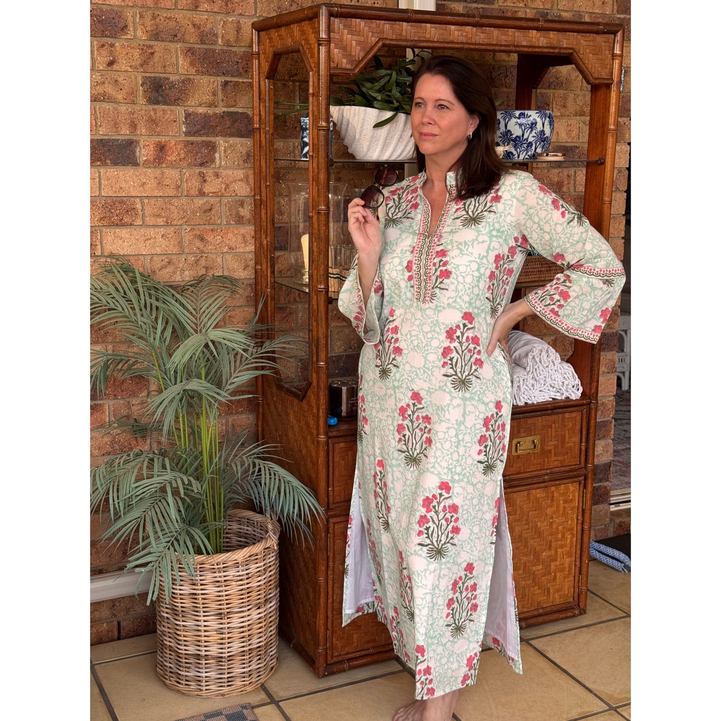 Woman in a floral dress standing in front of a wooden cabinet with a plant and wicker basket in the foreground.