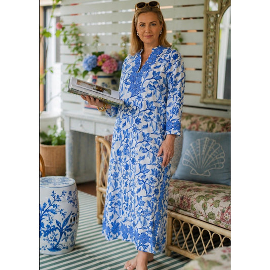 A woman wearing a blue and white floral print maxi dress stands in a decorated indoor setting with a bench, pillows, and plants.